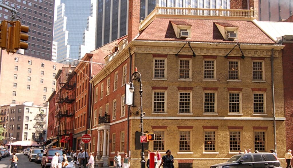Urban street with historic brick building, pedestrians, parked cars, traffic light, street signs.
