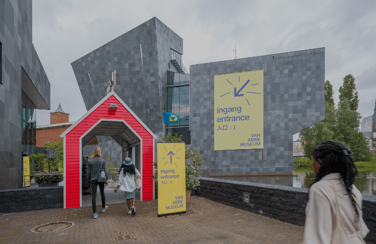Entrance to Van Abbe Museum with signs, modern gray buildings, red kiosk, and people walking.