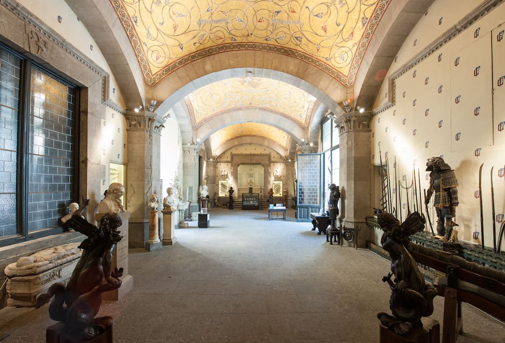 Vaulted gallery with sculptures, artifacts, intricate ceiling, large windows, and detailed decor.