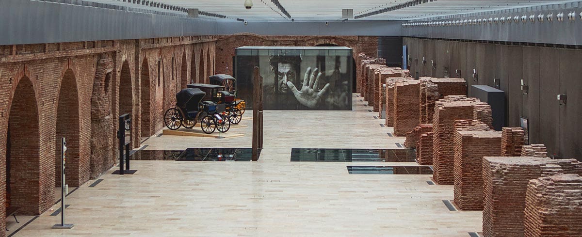Spacious indoor area with brick arches, vintage carriages, brick piles, and a large photograph.