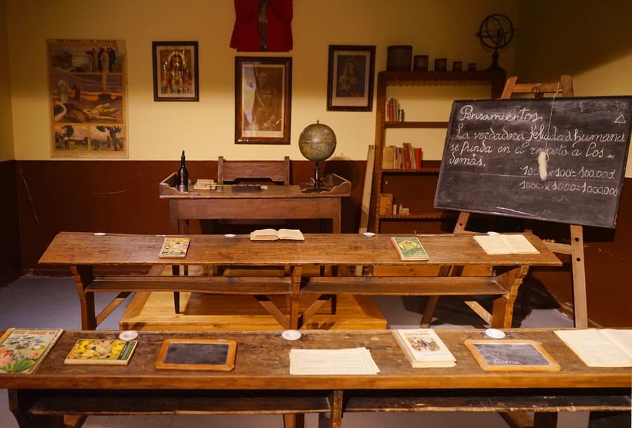 Vintage classroom with wooden desks, books, slate, teacher's desk, globe, chalkboard, and bookshelf.