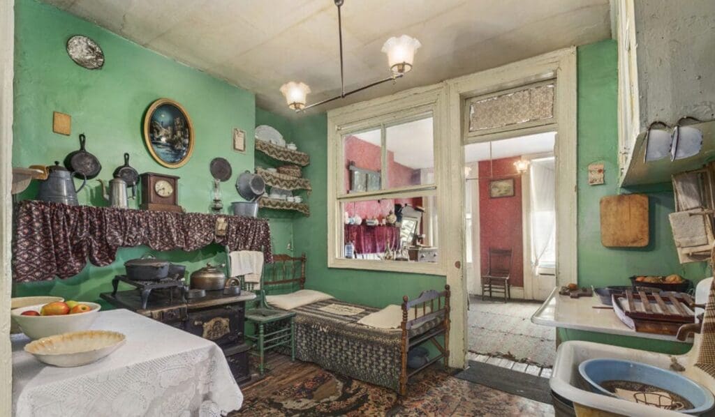 Vintage kitchen with green walls, historical kitchenware, a stove, dining table, bench, adjacent red room.