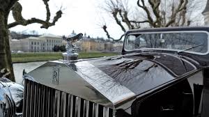 Vintage luxury car with prominent hood ornament, parked in a scenic area with trees and buildings.