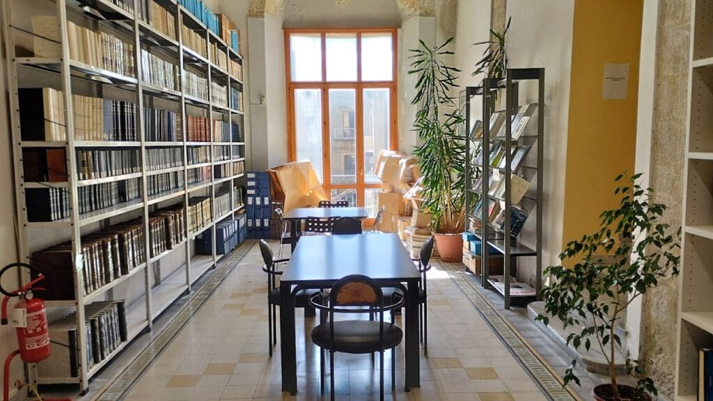 Well-lit library room with book-filled shelves, tables, and potted plants by large windows.