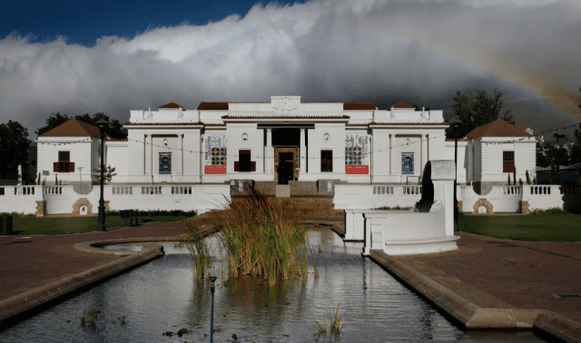 White building with columns, pathways, greenery, reflecting pool, statue, banners, and cloudy sky.
