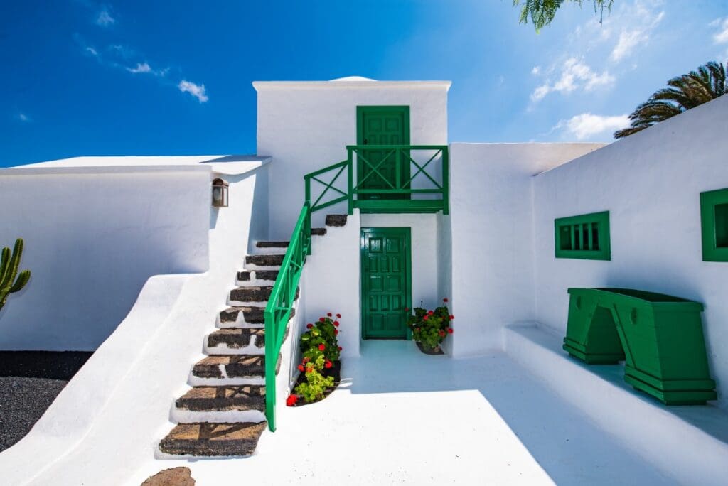 White building with green doors, windows, railings, potted red flowers, and an outdoor staircase.