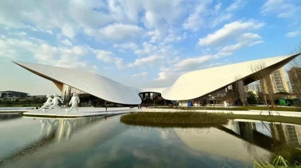 A curving, wing-like roofed building above a reflecting pool, surrounded by sculptures, trees, and grass.