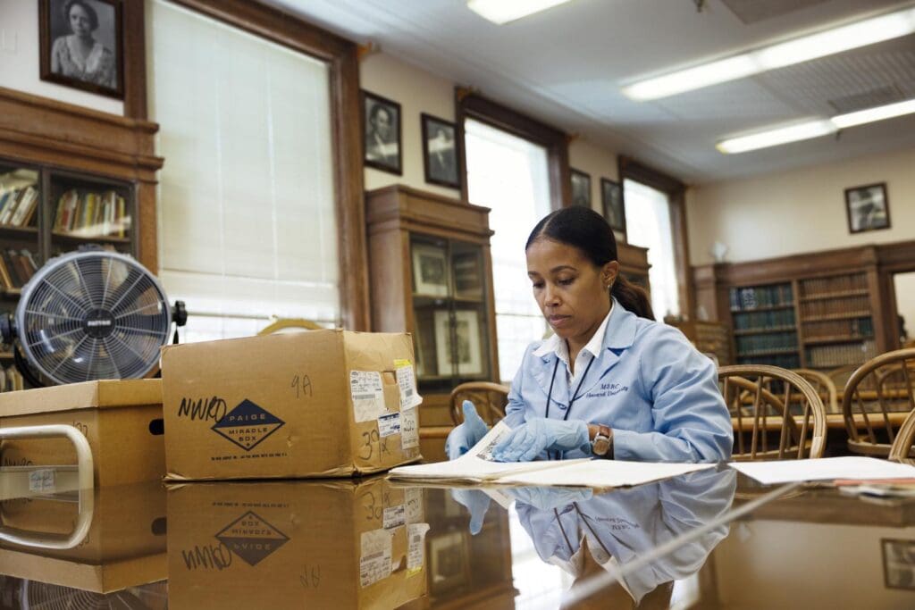 Woman in a blue lab coat examines documents at a wooden table in a library or archive room.