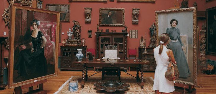A woman in a white dress views two large portraits in a warmly-colored art gallery filled with artworks.