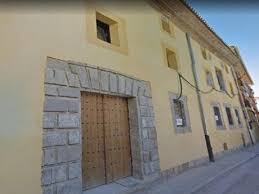 Yellow building with wooden door framed by stone blocks, windows with bars, along paved street.