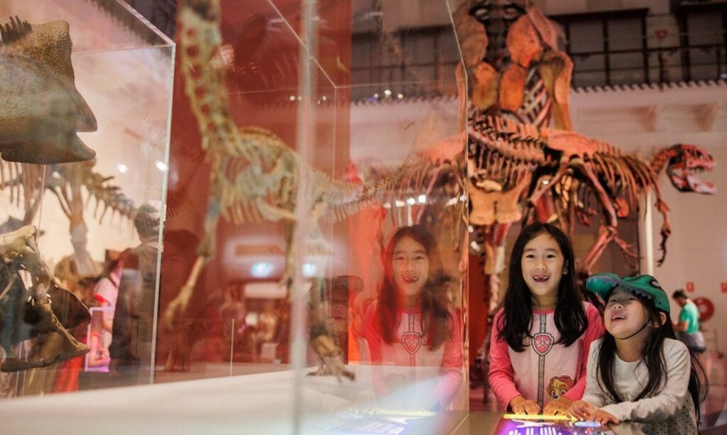 Children at a dinosaur exhibit with skeletons in the background at a museum.