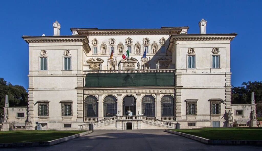 Grand building with ornate sculptures, balcony with flags, decorative facade, and well-kept grounds