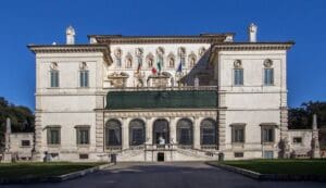 Grand building with ornate sculptures, balcony with flags, decorative facade, and well-kept grounds