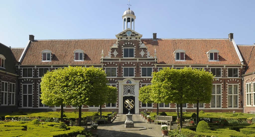 Historic brick building with red-tiled roof, central clock tower, and symmetrical garden pathways.