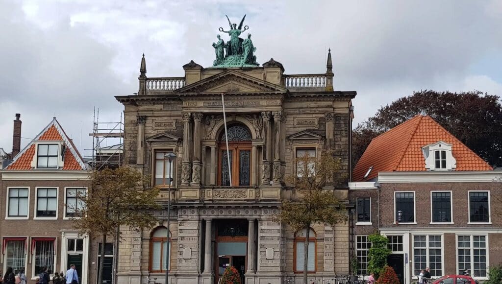 Historic stone building with columns, carvings, statues, flanked by Dutch buildings, cloudy sky.