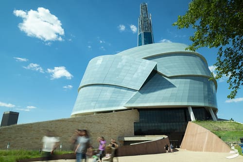 Modern circular architectural building with angular layers, clear sky, grassy landscaping, few people.