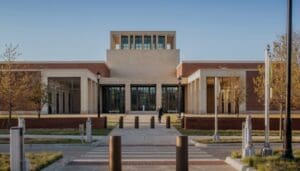 Modern institutional building exterior with brick and stone, trees, lampposts, and a path to glass doors.