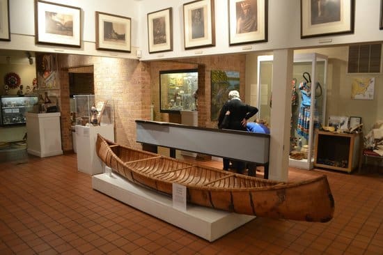 Museum room with wooden canoe on pedestal, framed art on walls, and glass display cases with artifacts.