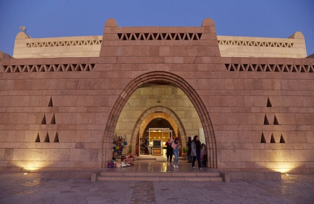 Large, stone building with arched entrance, geometric patterns, and people entering at twilight.