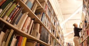 Woman organizes books on a tall library shelf, with another person browsing in the well-lit background.