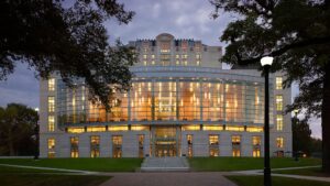 Modern multi-story building with arched glass facade, lit interior, and surrounding trees at twilight.