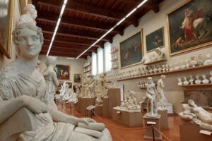 Museum gallery with classical sculptures, busts on pedestals, artwork on walls, high wooden ceiling.