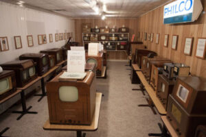 Room displaying a collection of vintage televisions with wooden walls and Philco sign on the right.