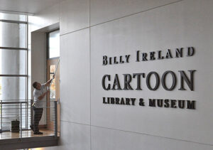 Person cleaning "Billy Ireland Cartoon Library & Museum" sign in modern, well-lit building.