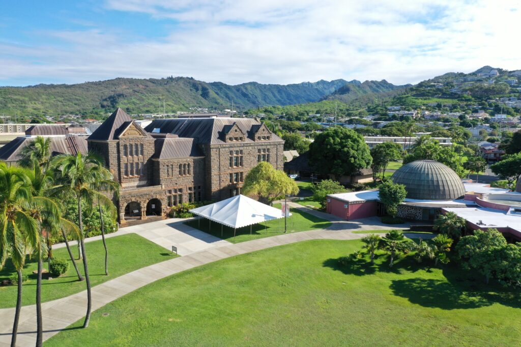 Historical stone building, domed structure, tent, trees, walkways, green lawn, mountains in background.