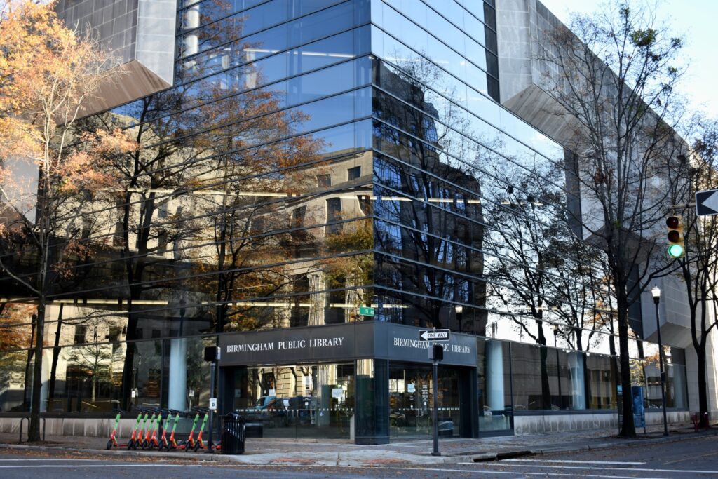Modern glass-walled Birmingham Public Library reflecting trees and urban surroundings, green traffic light.