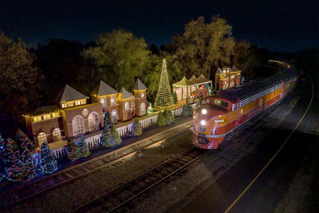 Festive vintage orange train passing illuminated holiday display with decorated houses and trees.