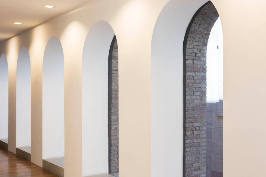 Hallway with arched openings, recessed ceiling lights, white walls, wooden floor, and brick wall view