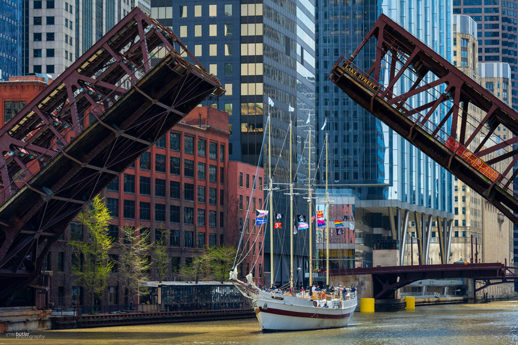 White sailboat with flags passing under a raised drawbridge in an urban area with high-rise buildings.
