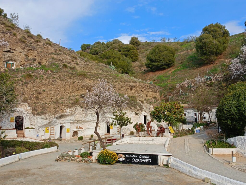 Hillside cave dwellings with Sacromonte Cave Museum sign, sparse vegetation, and clear skies.