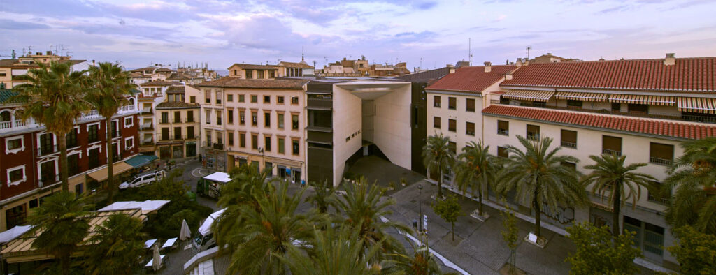 Open courtyard surrounded by multi-story buildings with red-tiled rooftops and scattered palm trees.