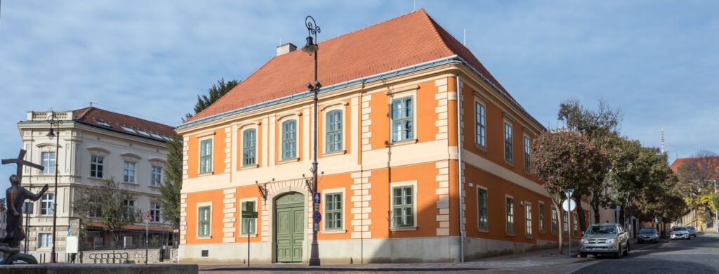 Historic building with orange facade, green shutters, and red roof on a quiet tree-lined street.
