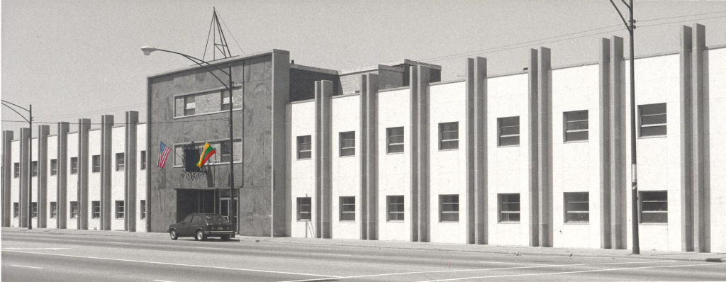 Black and white photo of a multi-story building with columns, two flags, and a parked car in front.