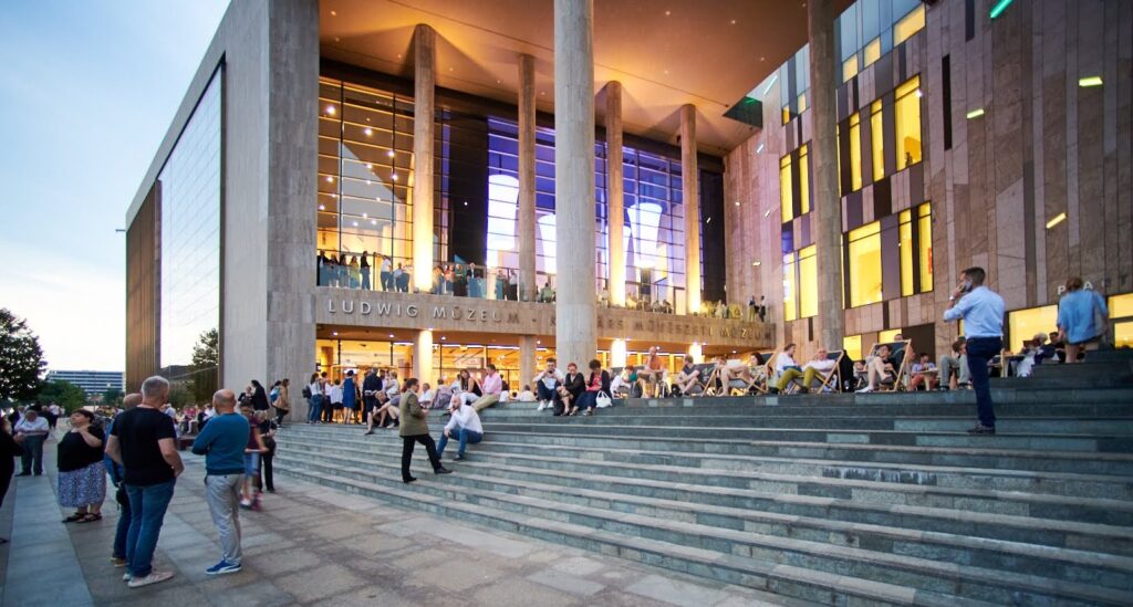 Modern building with large glass windows, tall pillars, people on wide steps; Ludwig Museum evening scene.