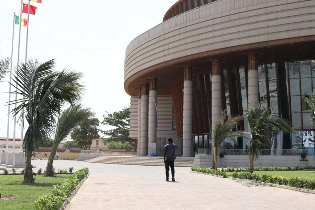 Man on pathway approaching a modern building with pillars, large windows, palms, and multiple flags.