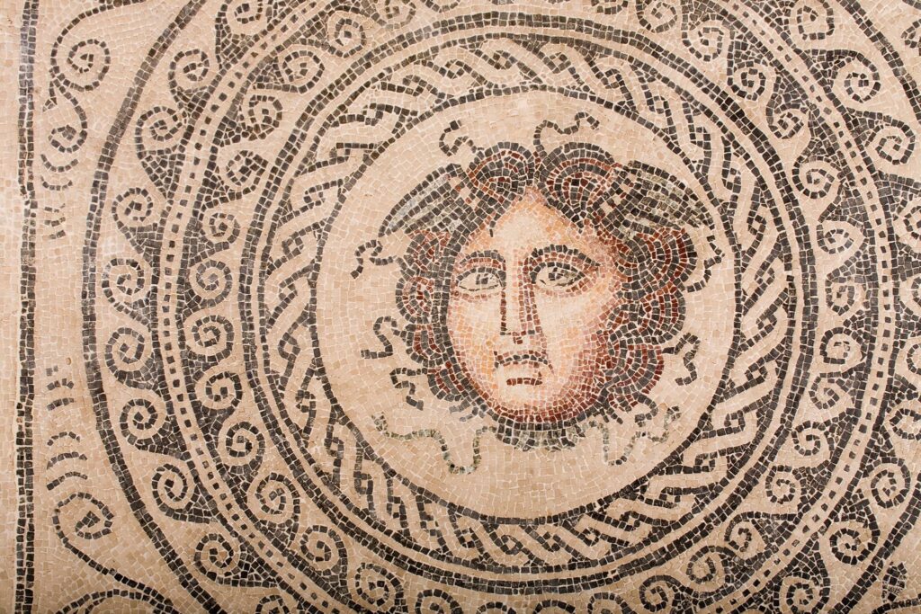 Mosaic artwork with a central face, geometric, and spiral patterns in earthy brown, black, and beige tones.