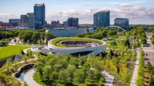 Modern urban landscape with a circular building, lush green park, high-rise buildings, and bridge.