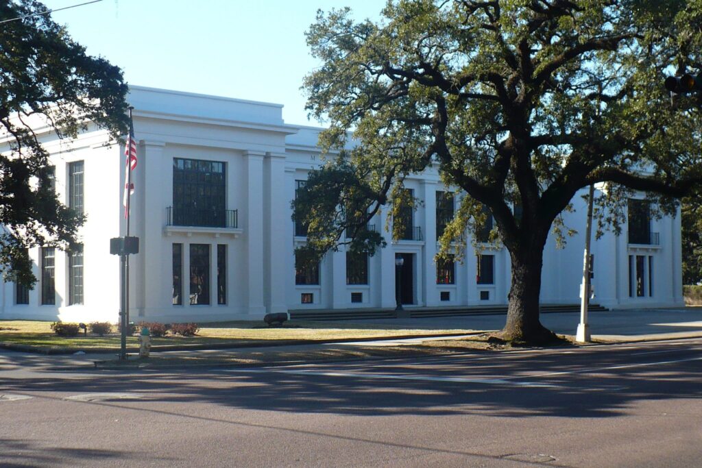 Large white classical building with columns and tall windows behind a tree, American flag in front.