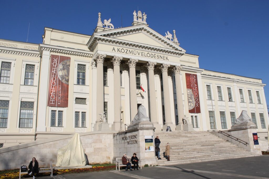 Móra Ferenc Museum, neoclassical building with columns, statues, banners, and people near entrance