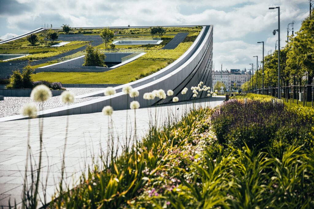 Modern sloped building with green roof, garden, flowers, lamp posts, and a city skyline in the distance.
