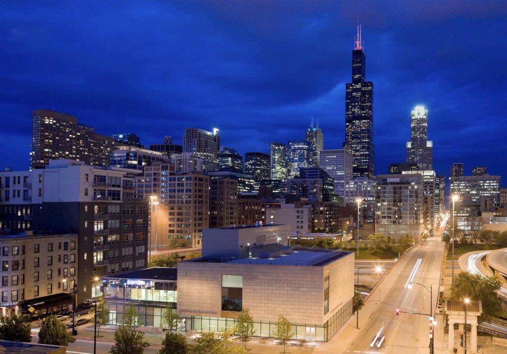 Nighttime cityscape with high-rise buildings, modern structure, roadway, and light trails.