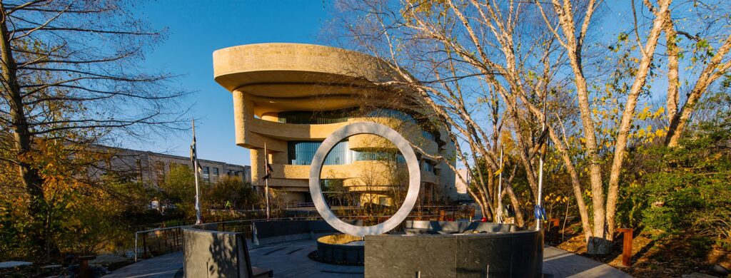 Curved modern beige building with glass windows, overhangs, trees, and circular sculpture in a plaza.