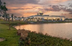 Modern building with a pyramid roof by water, surrounded by lawns, palm trees, and a sunset sky.