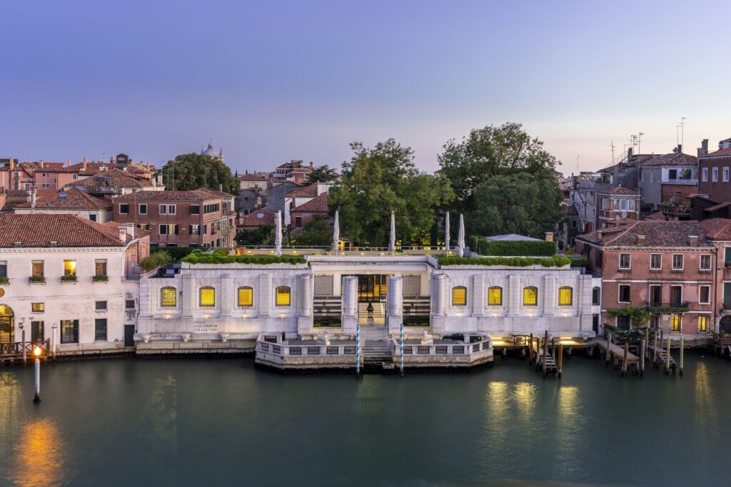 Historic waterfront building with classical architecture surrounded by red-roofed buildings and greenery.