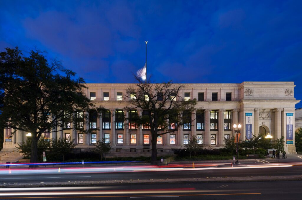 Neoclassical building at twilight with columns, archway, flying American flag, and light trails.