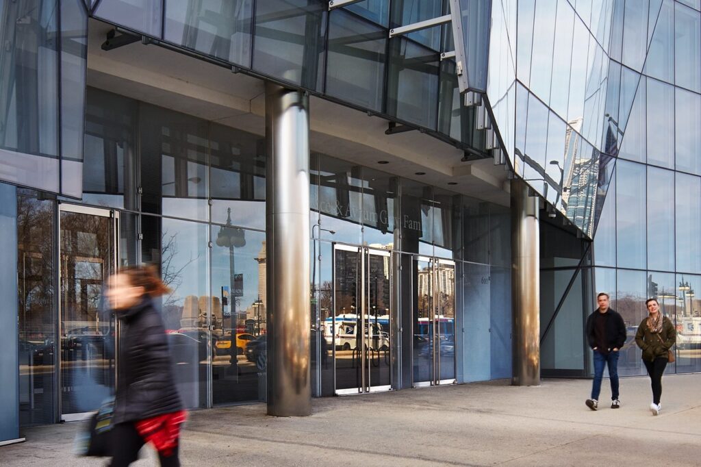 Modern glass building entrance with metallic columns and people walking by, reflections in windows.